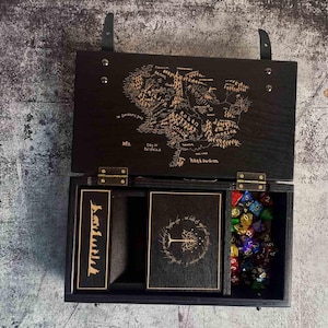 Può includere: Una scatola di legno nera con una mappa della Terra di Mezzo incisa sul coperchio. All'interno, un segnalibro, una carta con un disegno di un albero e una collezione di dadi poliedrici colorati. La scatola ha un cinturino in pelle e cerniere metalliche.