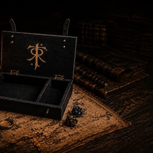 Op de afbeelding: Zwarte houten kist met een gouden embleem, open om twee compartimenten te onthullen. De kist staat op een kaart, met een kaars, dobbelstenen en een ring. De kist heeft een leren riem en is waarschijnlijk voor het opbergen van kleine items.