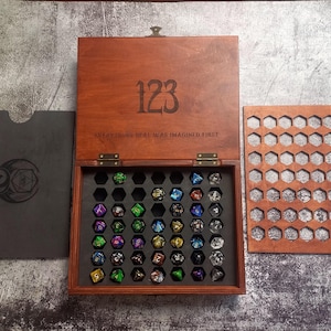 May include: Wooden dice box with a hinged lid, engraved with the number "123" and the quote "EVERYTHING REAL WAS IMAGINED FIRST." The box contains a variety of colorful polyhedral dice. A separate wooden tray with honeycomb cutouts is also visible.