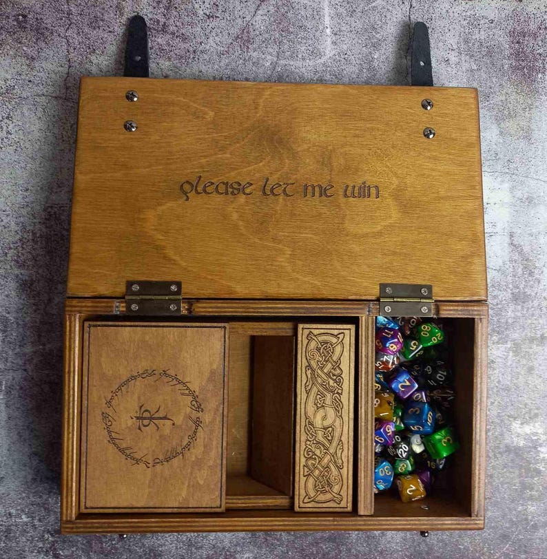 Op de afbeelding: Houten dobbelbox met compartimenten, met de tekst "please let me win" op het deksel. De doos bevat dobbelstenen, een kaart en een gesneden houten stuk. De doos is gemaakt van lichtbruin hout en heeft metalen scharnieren.