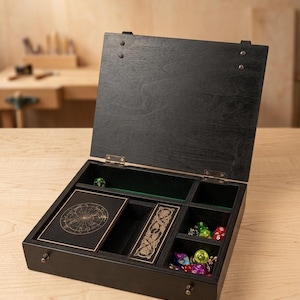 May include: A black wooden gaming box with an open hinged lid, featuring multiple interior compartments lined with green felt. The box contains a deck of cards with a celestial design, a rectangular card case with Celtic knotwork, and various colourful polyhedral dice.