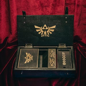 May include: A black wooden storage box with a hinged lid featuring a gold-coloured Triforce emblem. The interior contains three compartments, one holding a square card with a gold emblem, another a rectangular piece, and a third with dice icons.
