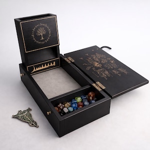 May include: Black wooden dice rolling box with a hinged lid and a separate dice storage compartment. The box features gold-coloured engraved designs and a metal leaf-shaped pin. The box contains various coloured dice.