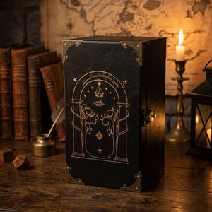 May include: A rectangular black box with a textured finish, featuring gold-coloured metal corner protectors and a central gold-toned latch. The front panel displays a gold-ink illustration of an arched doorway with trees, a crown, and text.