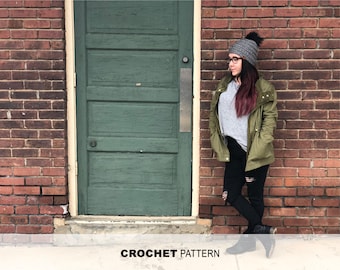May include: A woman wearing a gray sweater, a green jacket, black jeans, and a gray beanie with a white pom pom. She is standing in front of a brick wall with a green door.