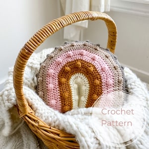 Può includere: Un cuscino arcobaleno all'uncinetto con una superficie testurizzata e colori rosa, marrone e bianco. Il cuscino è in un cesto di vimini con una coperta bianca.