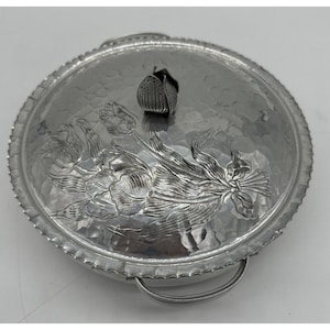 Puede incluir: Una fuente redonda plateada con tapa. La tapa presenta un diseño floral con un asa en forma de tulipán. La fuente tiene una textura martillada y un asa lateral. El diseño floral está en relieve en la tapa.