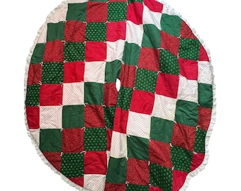 May include: A round Christmas tree skirt with a patchwork design in red, green, and white squares. The skirt has a ruffled white edge. The fabric squares feature solid colors and small patterns, creating a festive holiday look.
