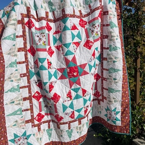 Peut inclure: Une courtepointe de Noël faite à la main avec un motif festif. La courtepointe présente un fond blanc avec des accents rouges, sarcelle et marron. La courtepointe a un motif d'étoile au centre et une bordure avec des arbres de Noël et des étoiles. La courtepointe est en tissu.