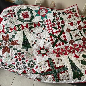 Peut inclure: Une courtepointe de Noël faite à la main avec un motif patchwork rouge, vert et blanc. La courtepointe présente divers motifs festifs, notamment des étoiles, des arbres et des formes géométriques. Elle a une bordure rouge et est probablement en coton.