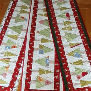 Peut inclure: Trois chemins de table sur le thème de Noël. Chaque chemin de table présente un fond blanc avec des formes d'arbres vertes et dorées, bordé de tissu rouge avec un motif festif. L'un a une bordure verte.