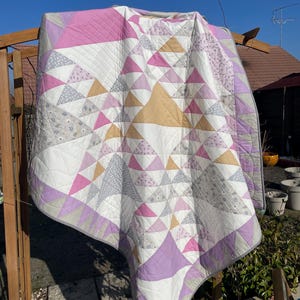 Peut inclure: Une courtepointe blanche, violette, rose et grise avec un motif géométrique de triangles. La courtepointe est suspendue à une structure en bois à l'extérieur.