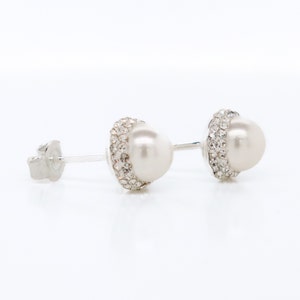 May include: A pair of silver stud earrings with a white pearl center and a clear crystal halo.