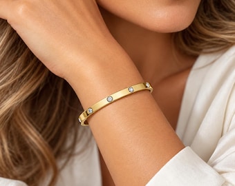 Puede incluir: Una pulsera rígida de tono dorado con una banda de metal lisa y pulida, engastada con varias piedras pequeñas, redondas y transparentes que brillan, distribuidas uniformemente por la superficie exterior de la pieza.