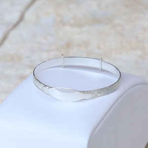 May include: A silver baby bangle with a blank oval-shaped tag for engraving. The bangle is decorated with a floral pattern.