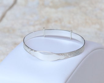Pode incluir: Um bracelete de bebé de prata com uma etiqueta oval em branco para gravar. O bracelete é decorado com um padrão floral.