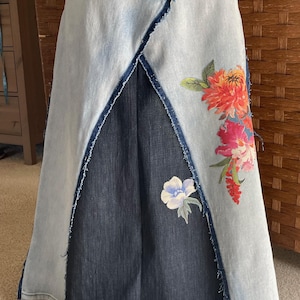 Puede incluir: Falda vaquera con un lavado azul claro y un panel azul oscuro en el centro. La falda presenta un diseño floral en tonos naranja, rosa y azul. Los bordes tienen un detalle deshilachado.