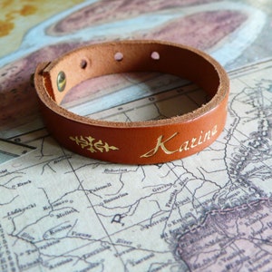 Peut inclure: Un bracelet en cuir marron avec un lettrage doré qui dit "Karine". Le bracelet a un motif floral décoratif.