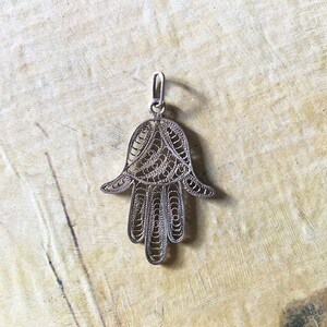 Può includere: Un ciondolo a forma di mano di Hamsa in filigrana d'argento. La mano di Hamsa è un simbolo popolare di protezione e buona fortuna in molte culture.