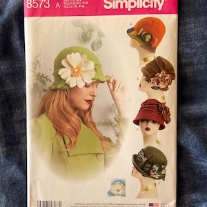 May include: Simplicity sewing pattern 8573 for hats in sizes S-L. The package features a large image of a green hat with a white flower and four smaller images of hats in orange, brown, red, and green with various floral and fabric embellishments.