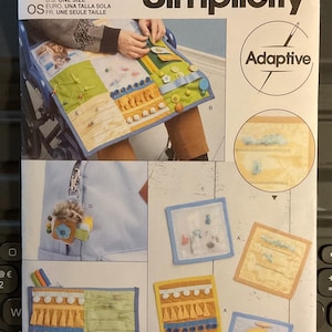 May include: Simplicity S9575 sewing pattern for an adaptive activity panel. The pattern includes various sensory elements, such as buttons, zippers, and textured fabrics. The image also shows a small keychain with a plush toy.