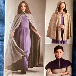 May include: A sewing pattern illustration featuring three cape styles. Style A is a long, floor-length grey cape. Style B is a shorter, hooded beige cape. Style C is a dark purple shoulder cape with a high collar, worn over a beige long-sleeve top.