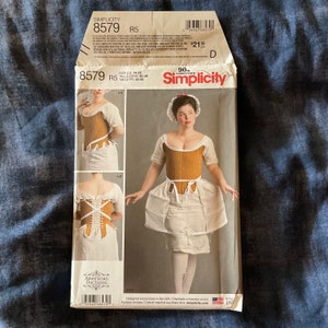 May include: A Simplicity sewing pattern for a corset, size US 14-22, Euro 40-48. The pattern shows a woman wearing a brown corset with a white skirt. The pattern is labeled "Simplicity 8579 R5".