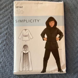 May include: A Simplicity costume pattern packet. The packet features a child wearing a black hooded costume. The pattern includes a tunic, pants, and a cape. The packet has the text "COSTUME" and "SIMPLICITY".