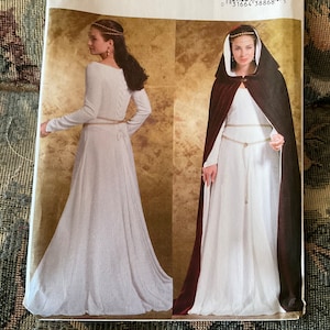 May include: A white dress with long sleeves and a fitted bodice. The dress has a lace-up back and a belt. The model is wearing a gold crown and a brown hooded cape.