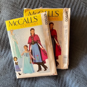 May include: Two sewing patterns for children's costumes. The pattern on the left is for a blue and white dress with a cape. The pattern on the right is for a red and black dress with a cape. Both patterns are from McCall's Costumes and are labeled M7000.