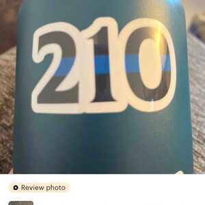 Puede incluir: Una botella de agua azul con una pegatina blanca con el n&uacute;mero "210" en negrita, con una fina l&iacute;nea azul. La pegatina es un homenaje a los agentes de polic&iacute;a.