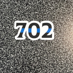 Puede incluir: Una pegatina blanca con el n&uacute;mero "702" en negro, contorneado en blanco. Una franja azul atraviesa horizontalmente el centro de los n&uacute;meros. La pegatina est&aacute; sobre una superficie gris moteada.