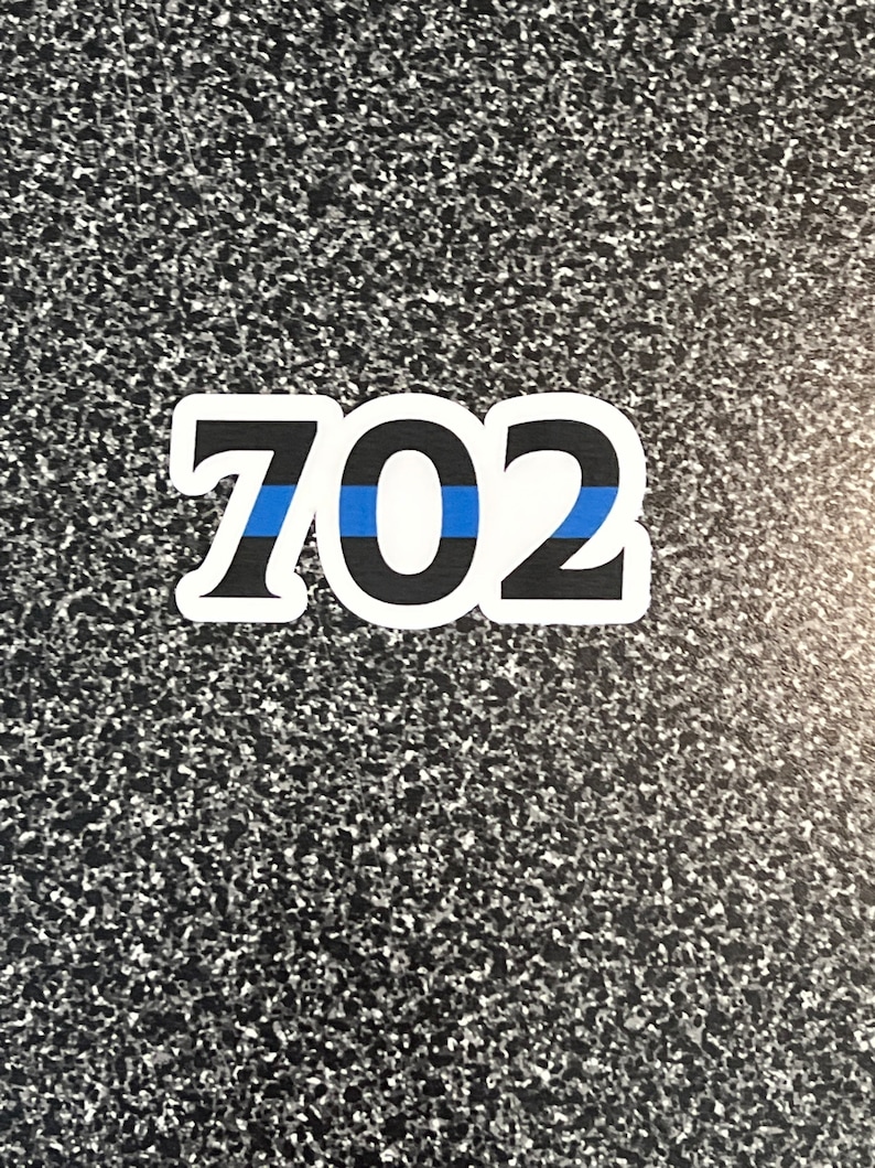 Puede incluir: Una pegatina blanca con el n&uacute;mero "702" en negro, contorneado en blanco. Una franja azul atraviesa horizontalmente el centro de los n&uacute;meros. La pegatina est&aacute; sobre una superficie gris moteada.