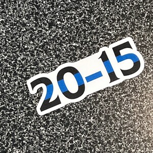 Puede incluir: Adhesivo blanco con el texto "20-15" en negro, con una franja azul. El adhesivo est&aacute; sobre una superficie moteada gris y negra.
