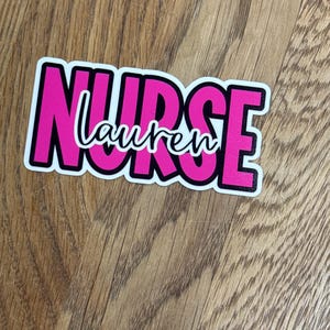 May include: A sticker with the word "NURSE" in large, bright pink letters with a black outline. The name "Lauren" is written in a cursive font in white letters inside the word "NURSE". The sticker is on a wooden surface.