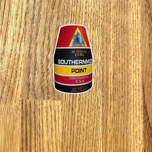 May include: A sticker featuring the Southernmost Point in Key West, Florida. The design includes a red top, black middle section, and yellow stripe, with text that reads "90 Miles to CUBA" and "CONTINENTAL U.S.A."