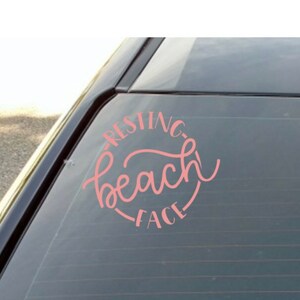 May include: A pink vinyl decal with the words "Resting Beach Face" in a circular design. The decal is applied to the rear window of a vehicle. The text is in a cursive font.