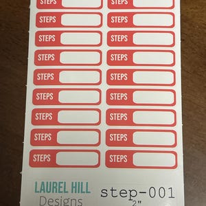 May include: A sheet of white stickers with red borders and the word "STEPS" printed on each. The stickers have a rectangular shape with a blank space for writing. The bottom of the sheet has the text "LAUREL HILL Designs" and "step-001 2"" printed on it.