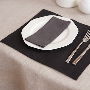 May include: A black placemat with a white plate, a grey linen napkin, a fork, and a knife. The placemat is on a beige linen tablecloth.