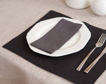 Op de afbeelding: Een zwarte placemat met een wit bord, een grijze linnen servet, een vork en een mes. De placemat ligt op een beige linnen tafelkleed.