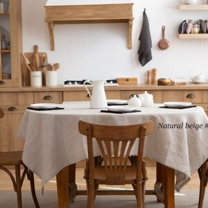 May include: A dining room scene featuring a wooden table set with a natural beige linen tablecloth. The table is set with white plates and black placemats. Wooden chairs surround the table. The kitchen has wooden cabinets and a white wall.
