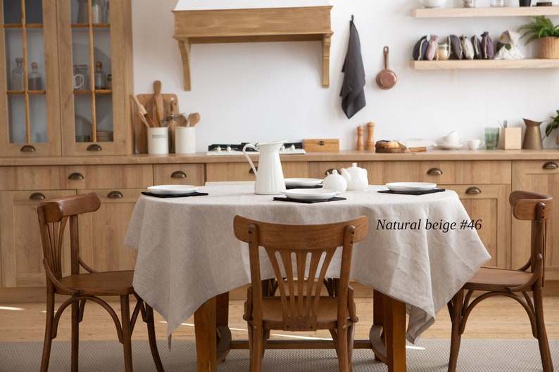May include: A dining room scene featuring a wooden table set with a natural beige linen tablecloth. The table is set with white plates and black placemats. Wooden chairs surround the table. The kitchen has wooden cabinets and a white wall.