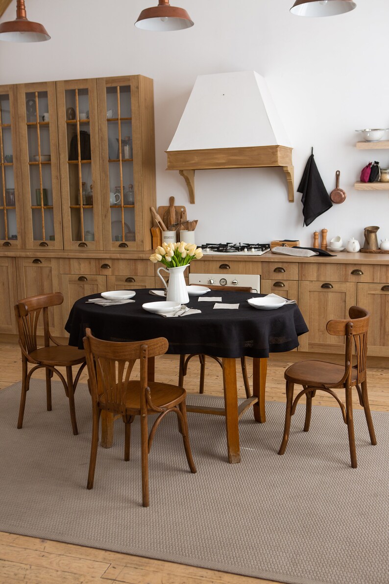 May include: A kitchen scene featuring a round wooden table set with white plates and a black tablecloth. Four wooden chairs surround the table. A white pitcher of yellow tulips sits in the center. The kitchen has wooden cabinets and a white range hood.