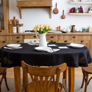 May include: A dining room scene featuring a wooden table set with a black tablecloth, white plates, and napkins. A white pitcher of yellow tulips sits in the center. Wooden chairs surround the table, and wooden cabinets and shelves are in the background.