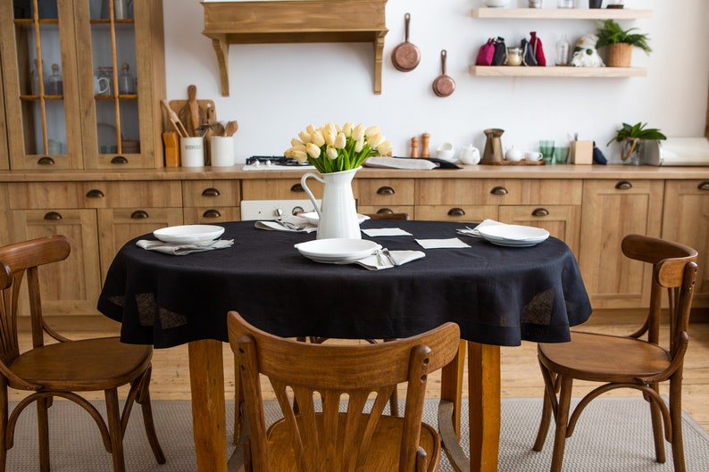 May include: A dining room scene featuring a wooden table set with a black tablecloth, white plates, and napkins. A white pitcher of yellow tulips sits in the center. Wooden chairs surround the table, and wooden cabinets and shelves are in the background.