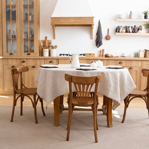 May include: A rustic dining room table setting with a light brown wooden table and four chairs. The table is covered with a beige linen tablecloth and has a white pitcher and black plates on top. The chairs are also made of light brown wood and have a simple design.