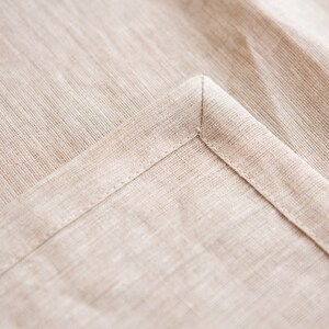 May include: Close-up of a beige linen fabric, showing a corner with a neatly sewn hem. The fabric has a subtle texture and a natural, neutral color. The stitching is precise, highlighting the quality of the textile.