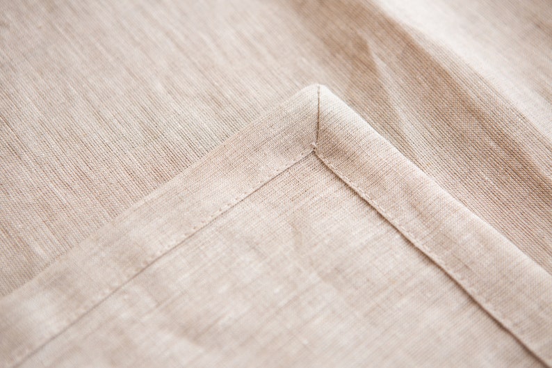 May include: Close-up of a beige linen fabric, showing a corner with a neatly sewn hem. The fabric has a subtle texture and a natural, neutral color. The stitching is precise, highlighting the quality of the textile.