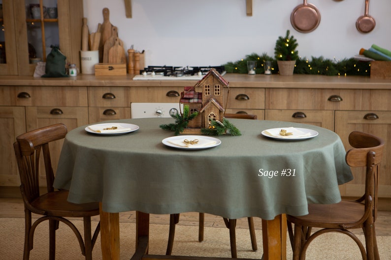 May include: A sage green round tablecloth on a wooden table, set with three white plates. A decorative wooden house and greenery are in the center. The text "Sage #31" is visible on the right side.