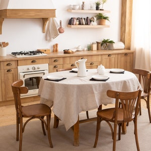 May include: A kitchen interior with wooden cabinets, a stove, and a white oven. A round table is set with a beige tablecloth, white plates, and black placemats. Four wooden chairs surround the table. A white pitcher and sugar bowl are on the table.